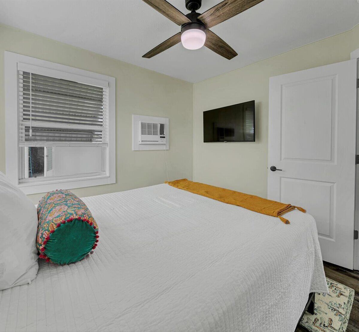 A bedroom with a bed, a ceiling fan, a window with blinds, a wall-mounted TV, and a door.
