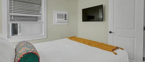 A bedroom with a bed, a ceiling fan, a window with blinds, a wall-mounted TV, and a door.