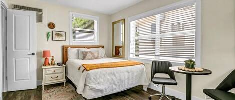 A bedroom with a bed, bedside table, lamp, chair, and a small table with a plant.