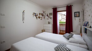 A bedroom with two beds, a dresser, a chair, and a window with red curtains. - Casa Shiva - Belvedere Dolomiti (Sedico)