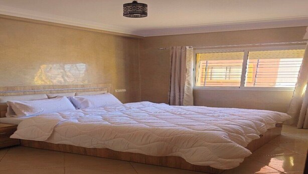 A bedroom with a large bed, a nightstand, a ceiling light, and a window with curtains. - Cosy apartment near the airport (Marrakech)