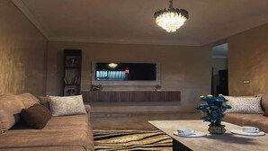 A modern living room with a sofa set, a coffee table with cups, a TV mounted on the wall, and a chandelier. - Cosy apartment near the airport (Marrakech)