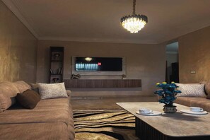 Ein modernes Wohnzimmer mit einer Sofagarnitur, einem Couchtisch mit Tassen, einem an der Wand montierten Fernseher und einer Kristallleuchtre.