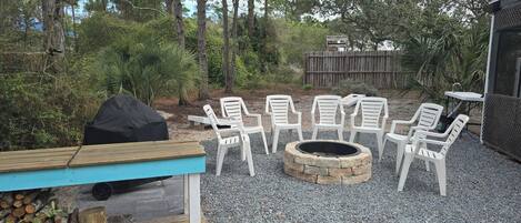 An outdoor seating area with white plastic chairs arranged around a stone fire pit, a wooden bench, and a grill.