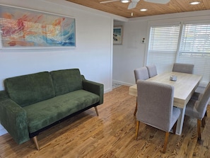 Une pièce avec un canapé vert, une table à manger en bois avec des chaises grises, un plancher en bois et un ventilateur de plafond.