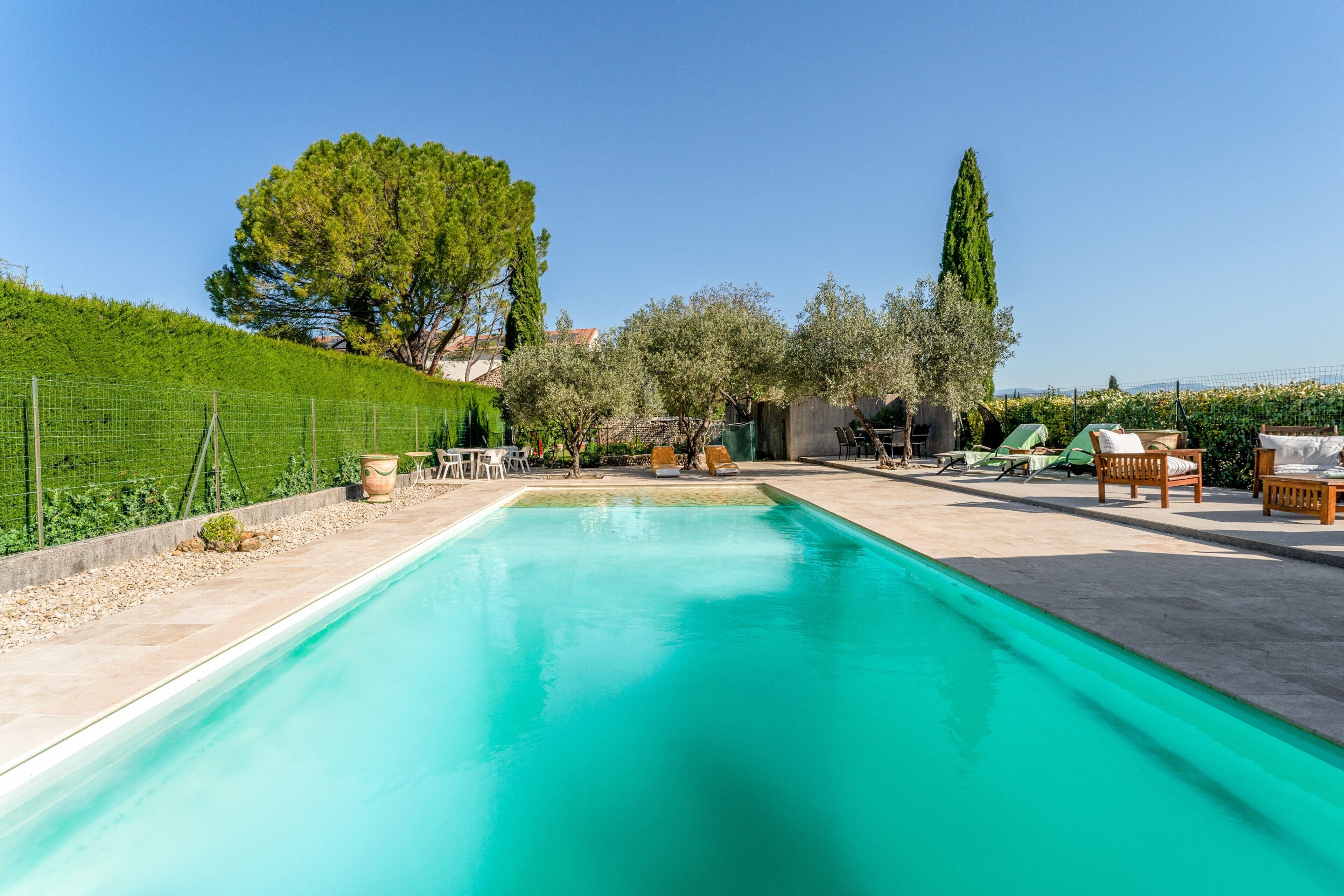 A swimming pool with a clear blue water, surrounded by a garden with trees and a fence.