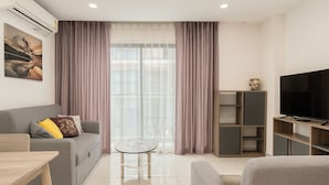 A modern living room with a grey sofa, a small round table, a TV on a wooden stand, and a large window with sheer curtains. - Cozy 2 Bedroom Condo in Pratumnak (Muang Pattaya)