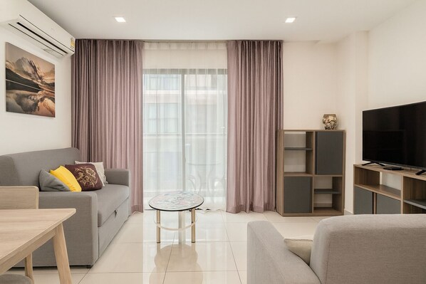 A modern living room with a grey sofa, a small round table, a TV on a wooden stand, and a large window with sheer curtains. - Cozy 2 Bedroom Condo in Pratumnak (Muang Pattaya)