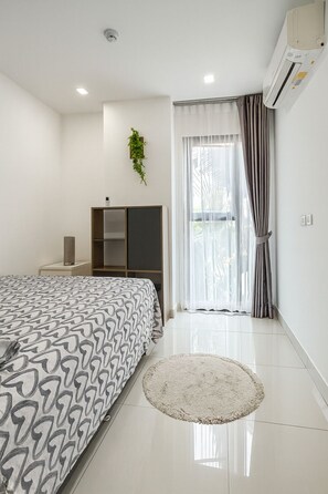 A modern bedroom with a large bed, a small bedside table, a built-in wardrobe, a round rug, and a window with curtains. - Cozy 2 Bedroom Condo in Pratumnak (Muang Pattaya)