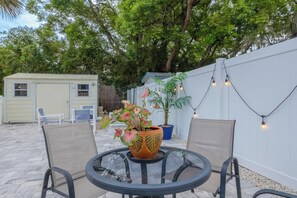 Un espace patio comprenant une table ronde en verre, deux chaises, une plante en pot et une guirlande lumineuse.