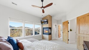 A bedroom with a bed, a fireplace, a ceiling fan, and large windows offering a view of the outdoors. - HotTub+Sauna+Garage+DeerValleyEast+Jordanelle (Heber City)