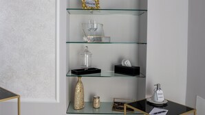 Un interior moderno con una estantería de vidrio que exhibe objetos decorativos, una mesa negra y dorada, y un piso de herringbone gris.