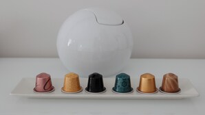 Un objeto blanco en forma de huevo sobre una superficie blanca, con un plato blanco debajo que contiene seis tazas de colores y diseños variados.