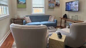 Una sala de estar con un sofá azul, dos sillones, una mesa de centro y un televisor montado en la pared.