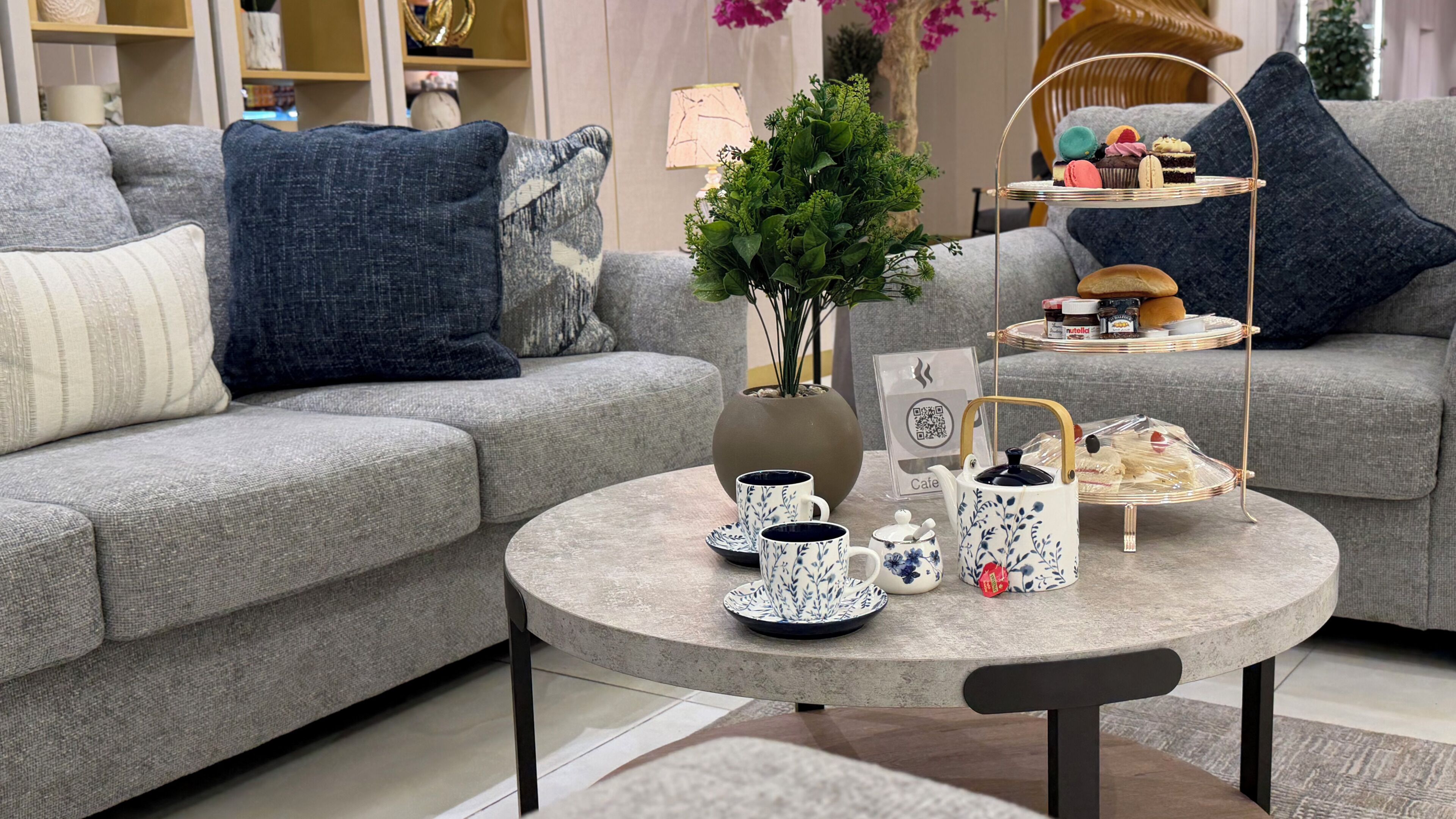 Un salon moderne avec un canapé gris, une table basse ronde et des objets décoratifs, notamment un vase avec des plantes vertes, un plateau de pâtisseries et une théière.
