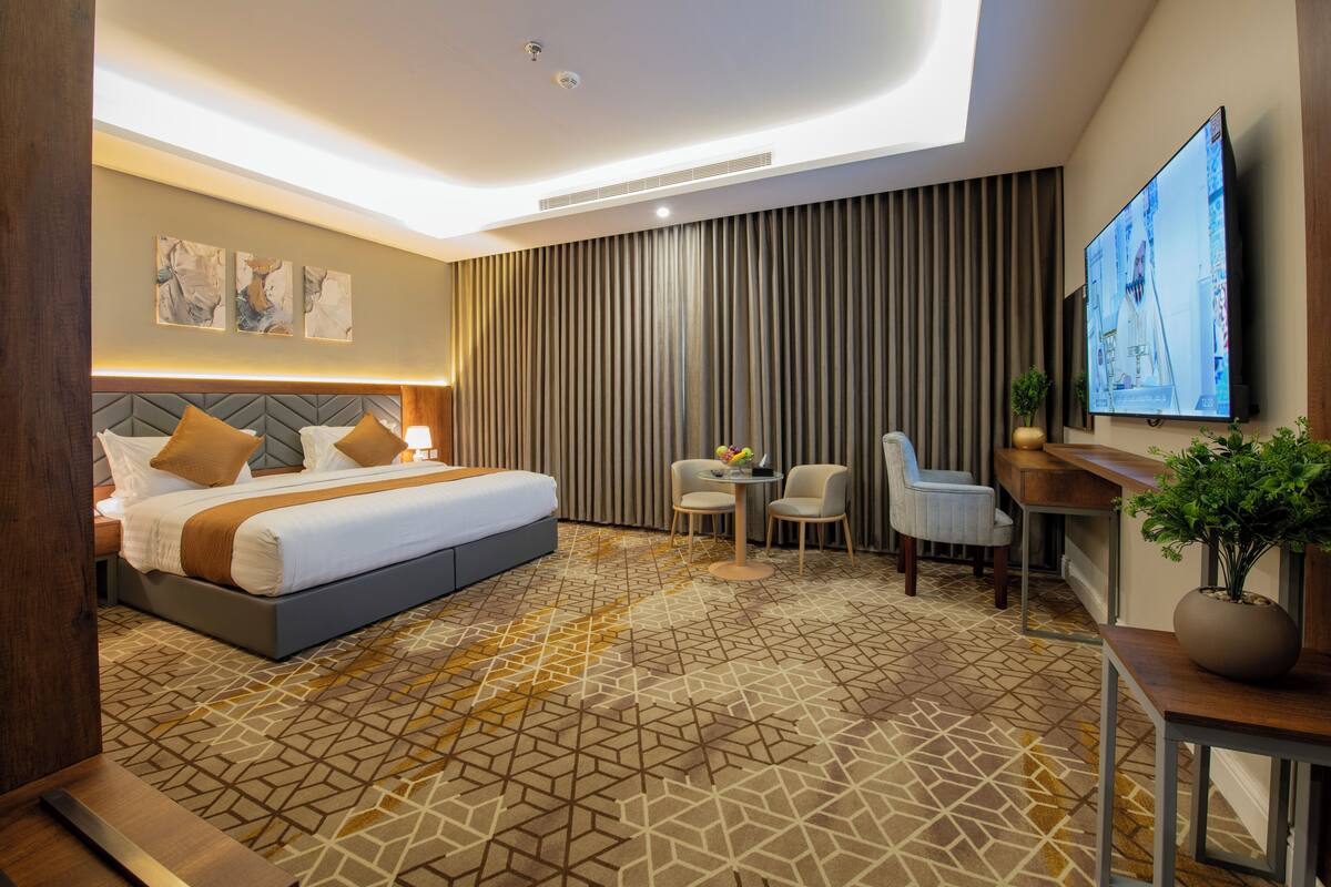 A modern hotel room with a large bed, a desk, a chair, a TV, and a patterned carpet.
