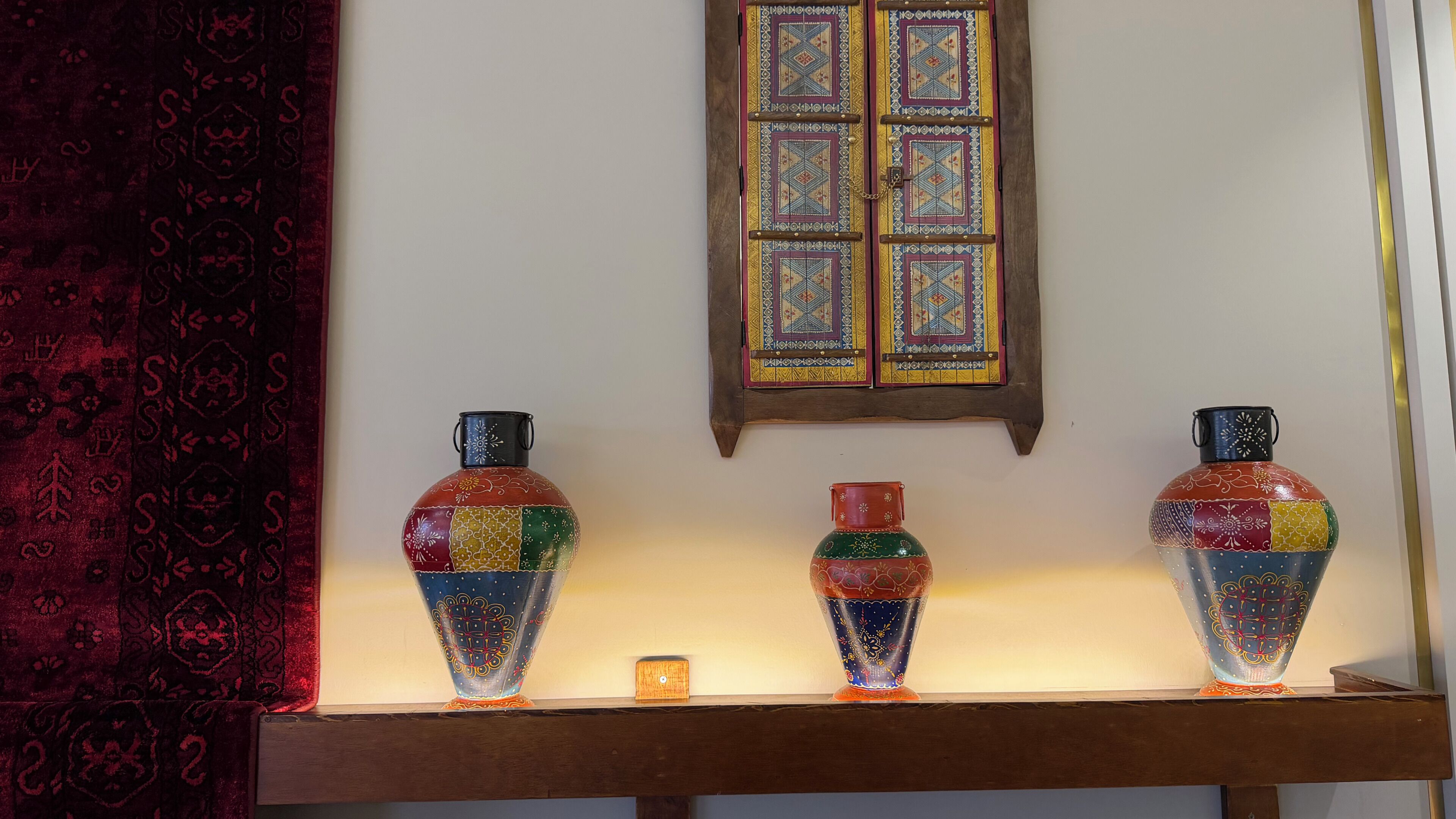 A display of colorful vases on a shelf with a patterned curtain and a window with a geometric design.