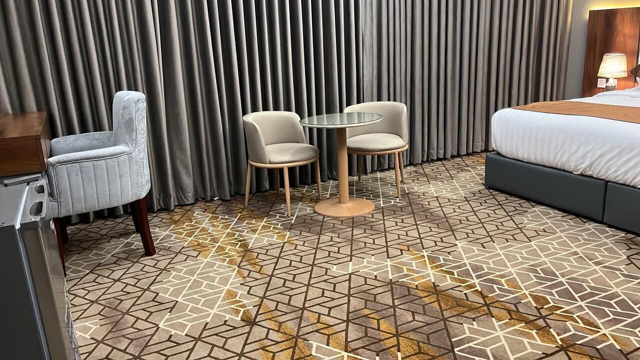 A hotel room with a bed, two chairs, a small table, and a patterned floor.