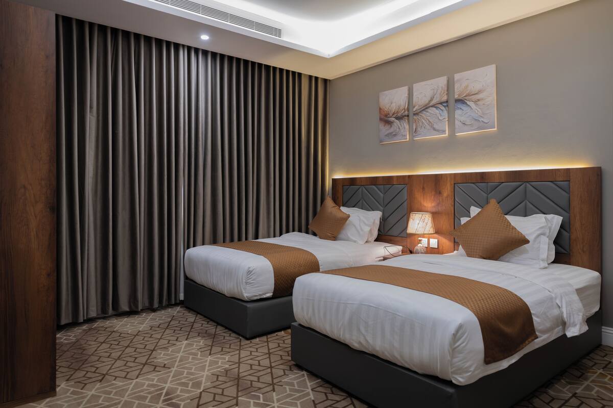 A hotel room with two beds, a large window with curtains, a wall-mounted air conditioner, and a geometric patterned floor.
