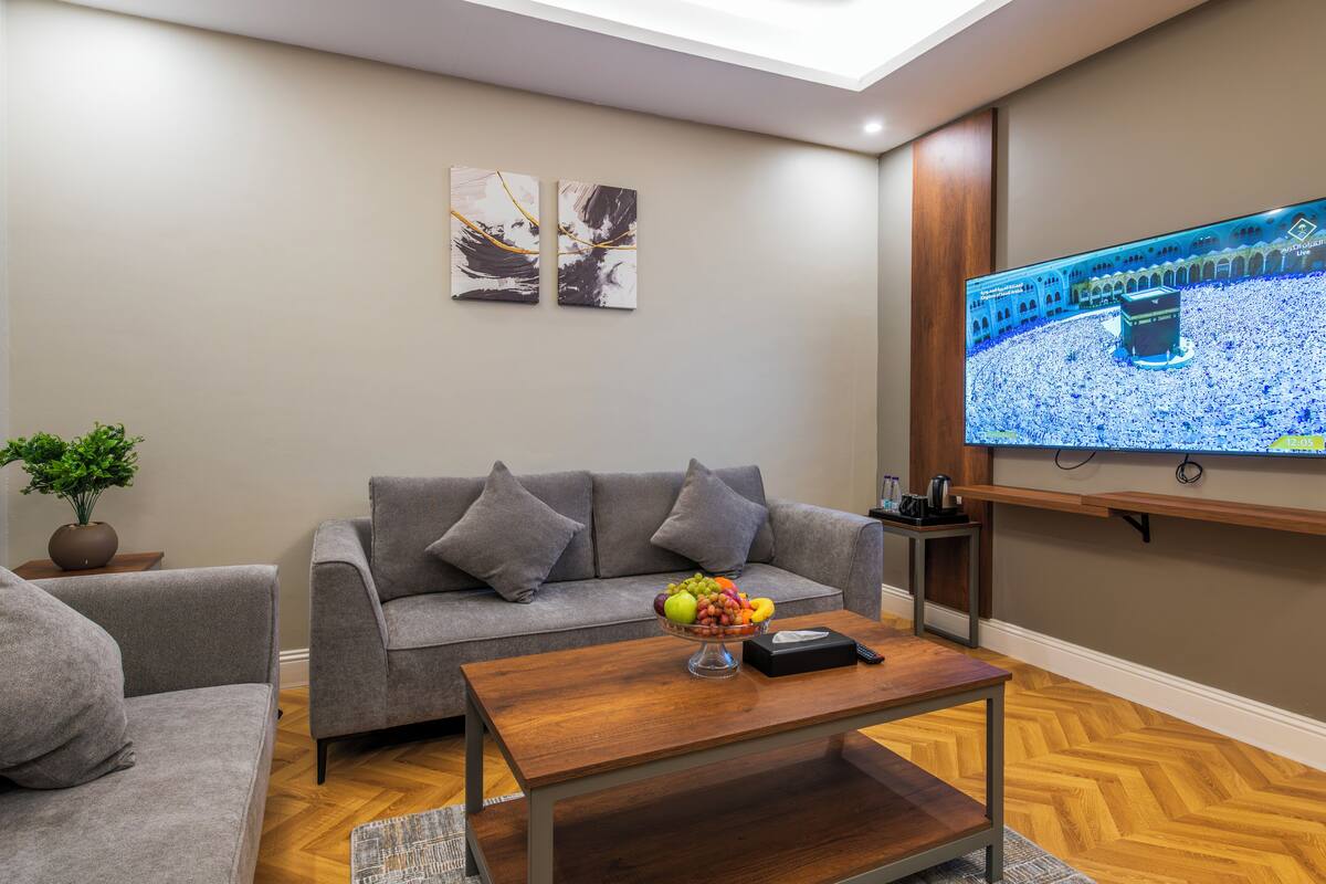 A modern living room with a grey sofa, a wooden coffee table with fruit, a flat-screen TV, and abstract wall art.