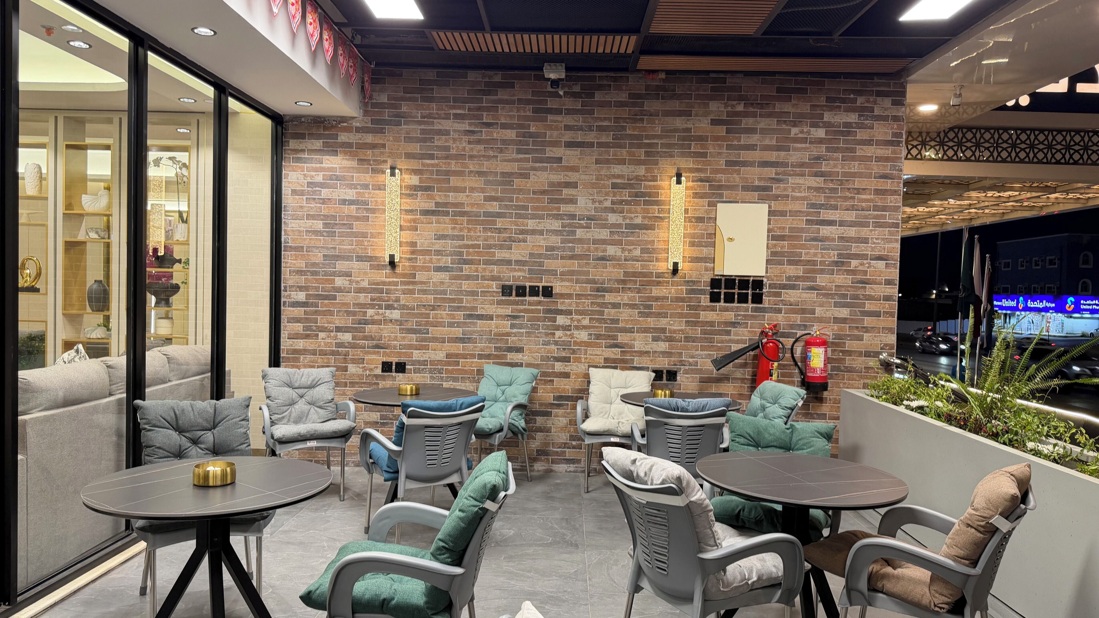A brick wall with a fire extinguisher, outdoor seating with cushioned chairs and tables, and a view of a street at night.