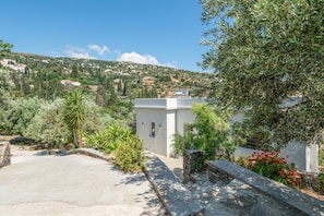 A modern building with a paved driveway, surrounded by greenery and a hillside view.