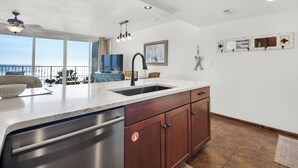 A modern kitchen with a large island, stainless steel dishwasher, wooden cabinets, and a view to the outside. - Beachfront front Condo with Stunning Views & Last-Minute August Discounts! (Panama City Beach)