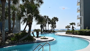 A swimming pool with a curved design, surrounded by palm trees and a building with balconies. - Beachfront front Condo with Stunning Views & Last-Minute August Discounts! (Panama City Beach)