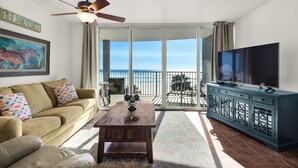 A living room with a sofa, coffee table, TV, and a balcony with a view of the ocean. - Beachfront front Condo with Stunning Views & Last-Minute August Discounts! (Panama City Beach)