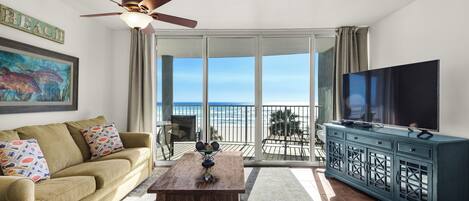 A living room with a sofa, coffee table, TV, and a balcony with a view of the ocean.