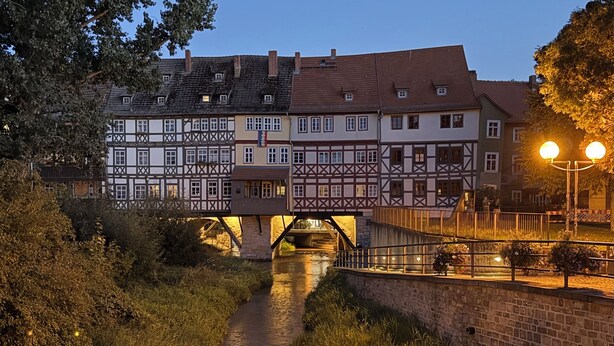 A traditional half-timbered building with a bridge over a narrow waterway, surrounded by greenery and street lamps. - Our new and cozy Krämernest in the old town, 100m to the Krämerbrücke (Erfurt)