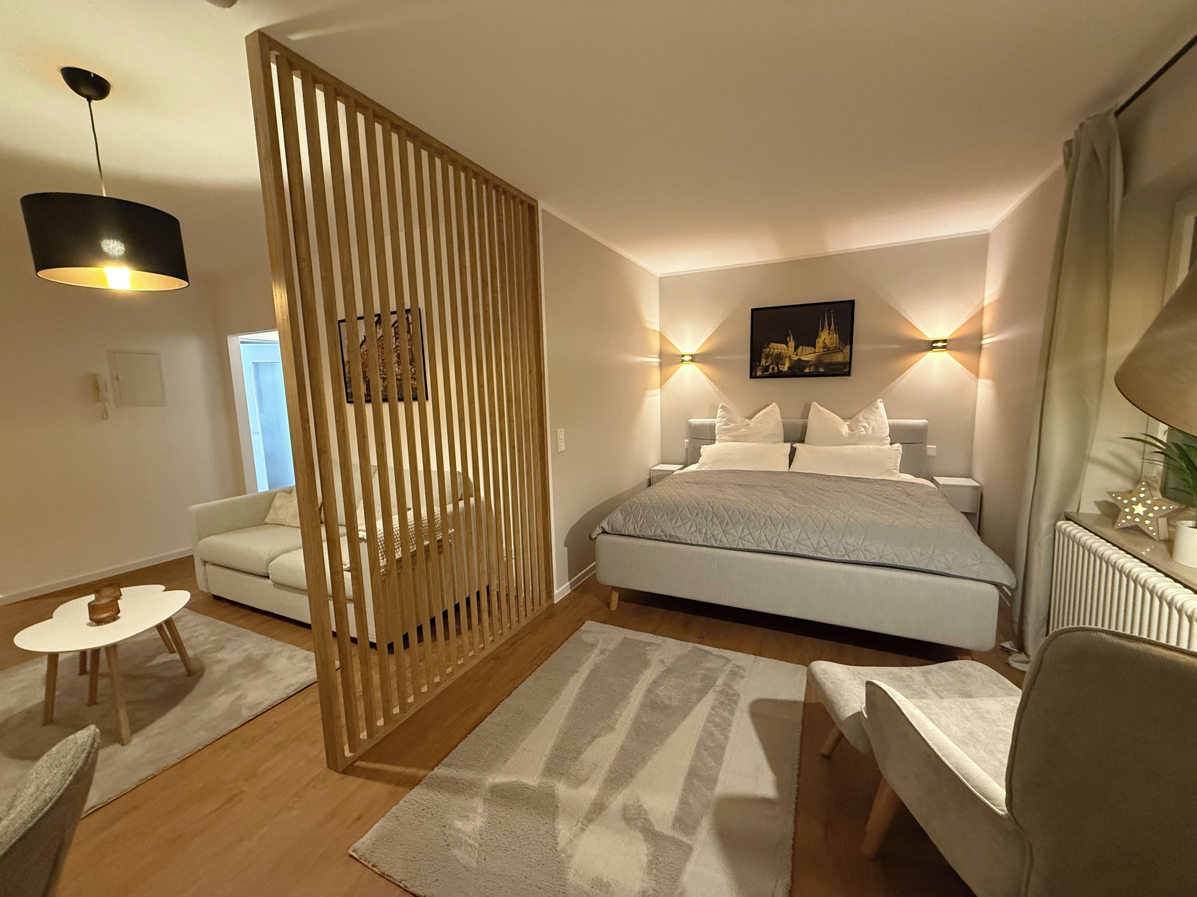 Ein modernes Schlafzimmer mit Bett, Nachttischen, einem Sessel, einem Schreibtisch, einem Dekosparren und einem Bild an der Wand.
