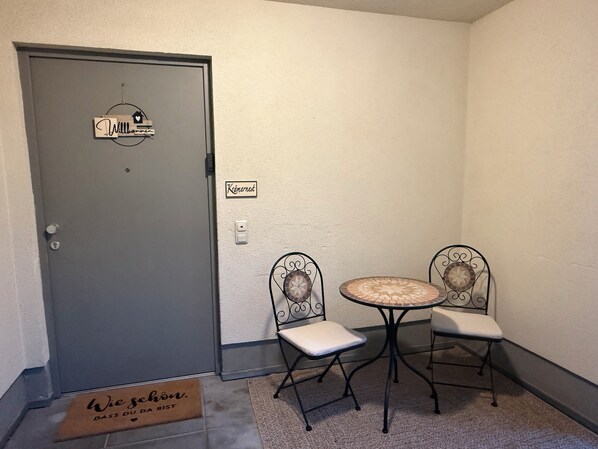 Une petite pièce avec une porte grise, une table ronde avec deux chaises, un paillasson et un interrupteur fixé au mur.