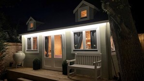 A small, well-lit house with a porch, a bench, and a large vase. - Cosy Alpine Lodge in Claygate (Claygate)