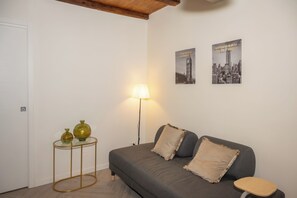 Una habitación con un sofá gris, dos almohadas, una mesita redonda con dos jarrones, una lámpara de pie y dos carteles en la pared.