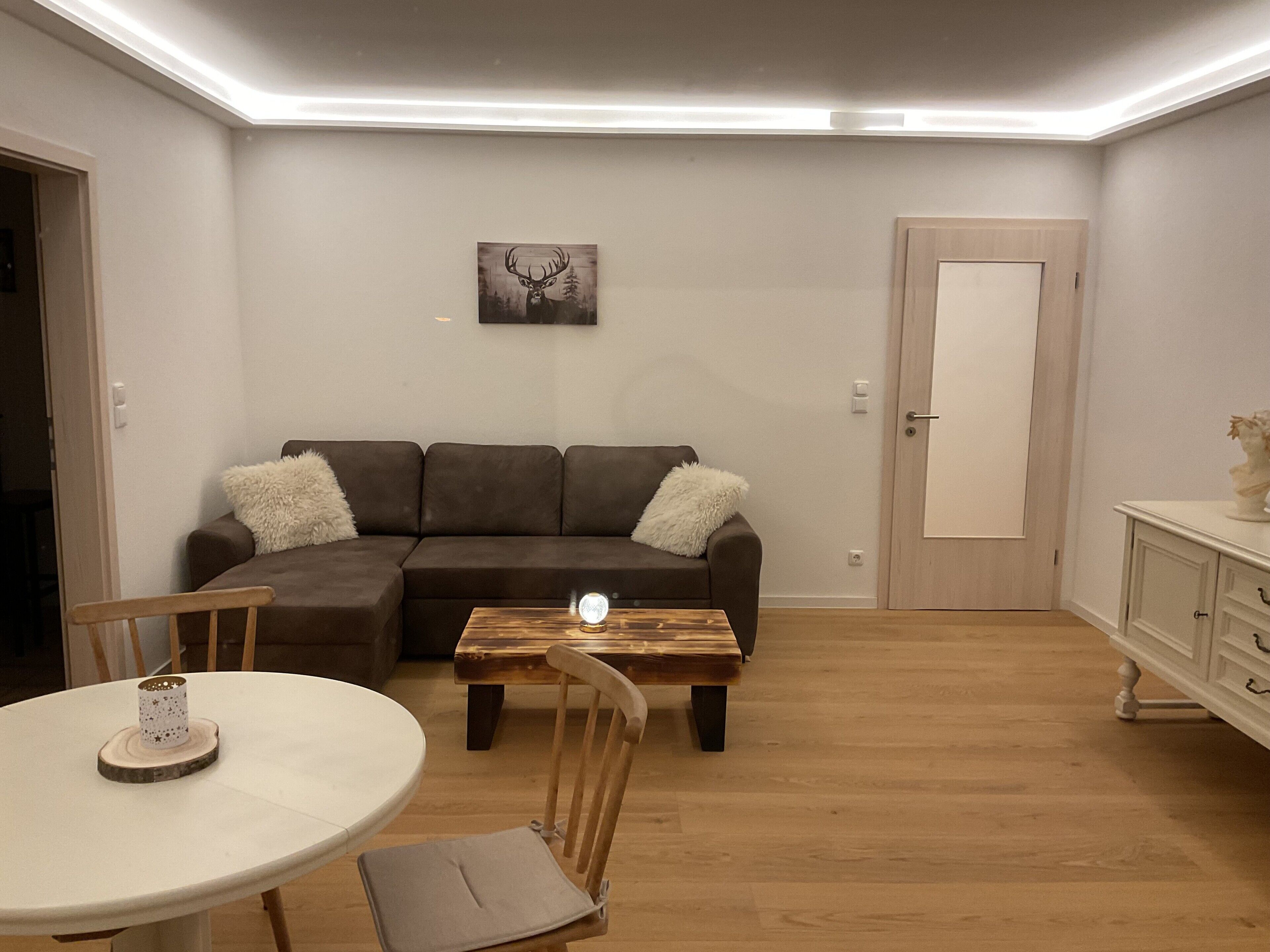 A living room with a brown sectional sofa, a wooden coffee table, a white round table with a chair, and a door with a white cabinet.