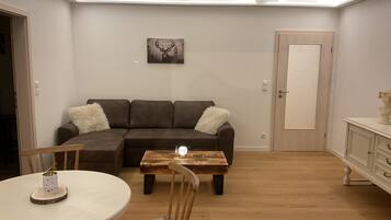 A living room with a brown sectional sofa, a wooden coffee table, a white round table with a chair, and a door with a white cabinet.