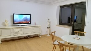 Una sala de estar con televisor, un mueble blanco, una estatua, una mesa redonda y sillas de madera.