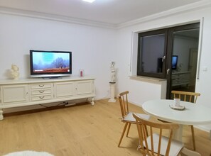 Una sala de estar con televisor, un mueble blanco, una estatua, una mesa redonda y sillas de madera.
