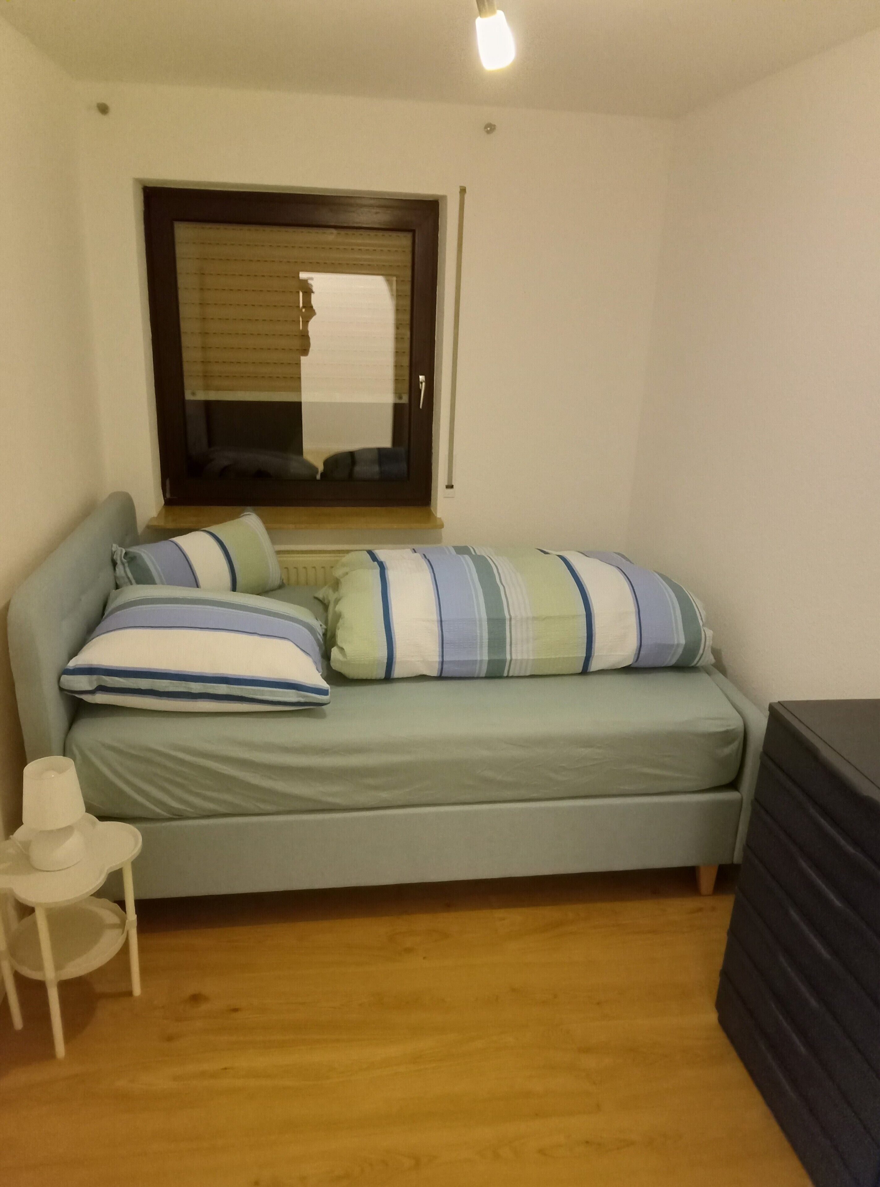 Une petite pièce comprenant un lit simple, une chaise, une commode et une fenêtre avec des stores.