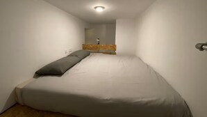 Ein kleiner, schmaler Raum mit einem Einzelbett, einem Holz-Kopfteil und einer Deckenleuchte.