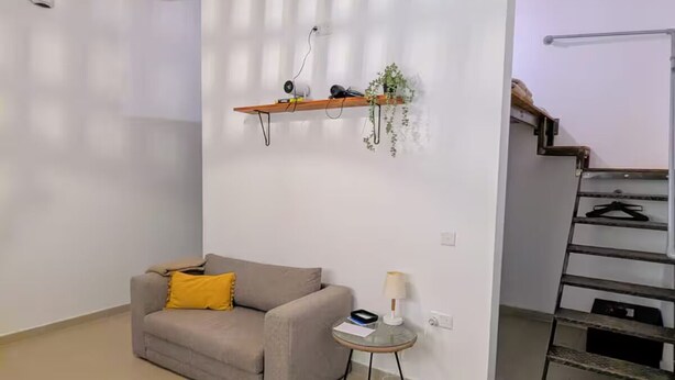 Ein modernes Wohnzimmer mit einer Couch, einem kleinen Tisch, einer Hängeleuchte, einem an der Wand befestigten Regal mit Pflanzen und einer Treppe.