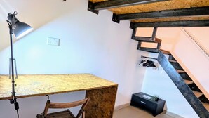 Ein Schlafzimmer im Loftsstil mit schrägem Dach, sichtbaren Holzbalken, einem Schreibtisch mit Stuhl, einer Treppe und einem kleinen schwarzen Schrank.