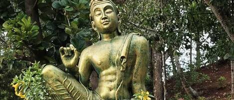 A seated statue of a figure in a natural setting with trees and plants around.