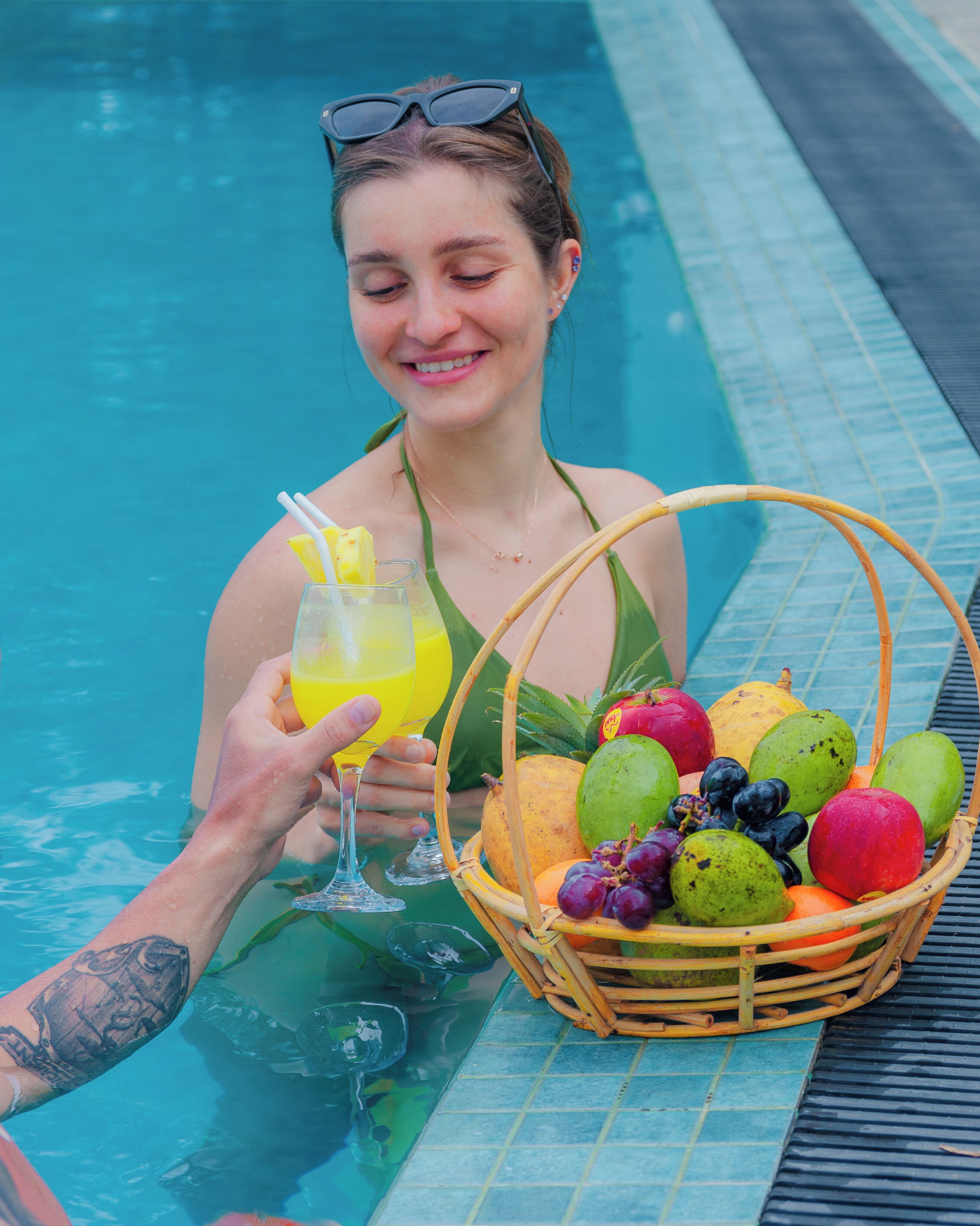 A person holding a glass of yellow drink and a basket of fruits by a pool.