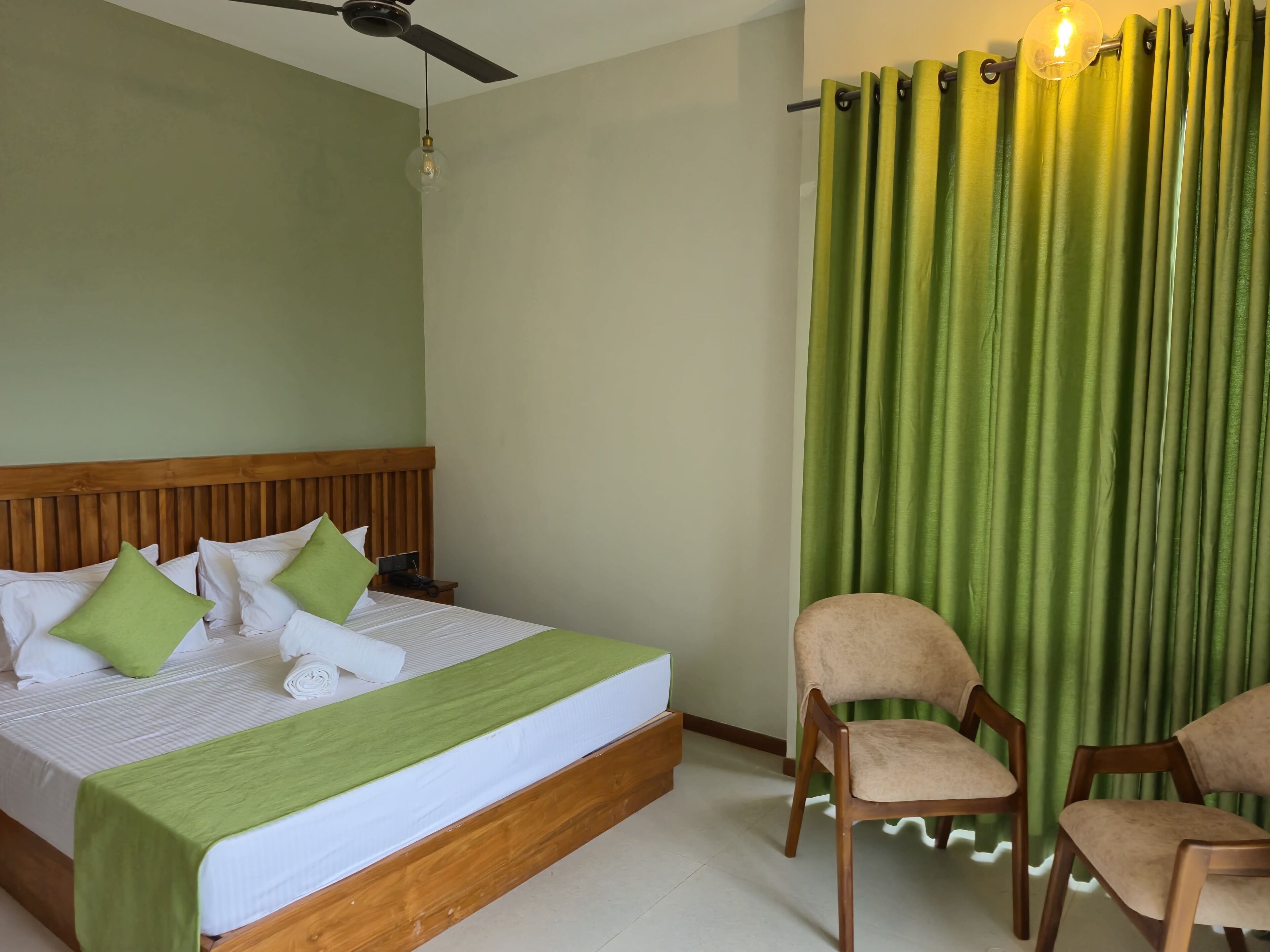 Un lit bien fait, avec une tête de lit en bois, deux oreillers verts et une couverture verte. Une chaise avec un coussin beige est placée à côté du lit, et un rideau vert recouvre partiellement une fenêtre.