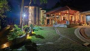 A well-lit outdoor area with a covered seating area, a pathway leading to a building, and a landscaped garden. - River Shades Hotel (Hikkaduwa)