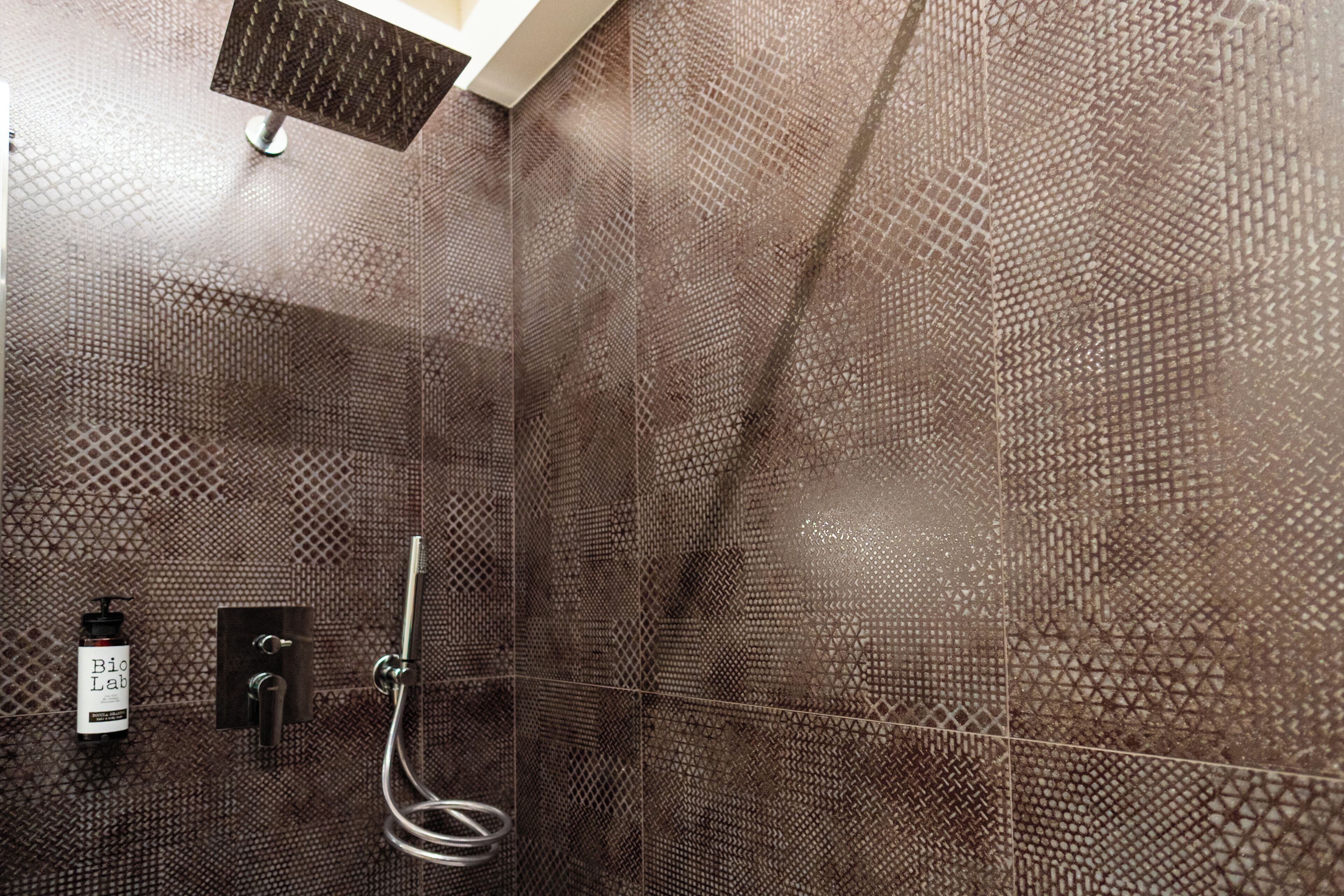 A shower with a geometric tile pattern, a handheld showerhead, and a bottle of shampoo.