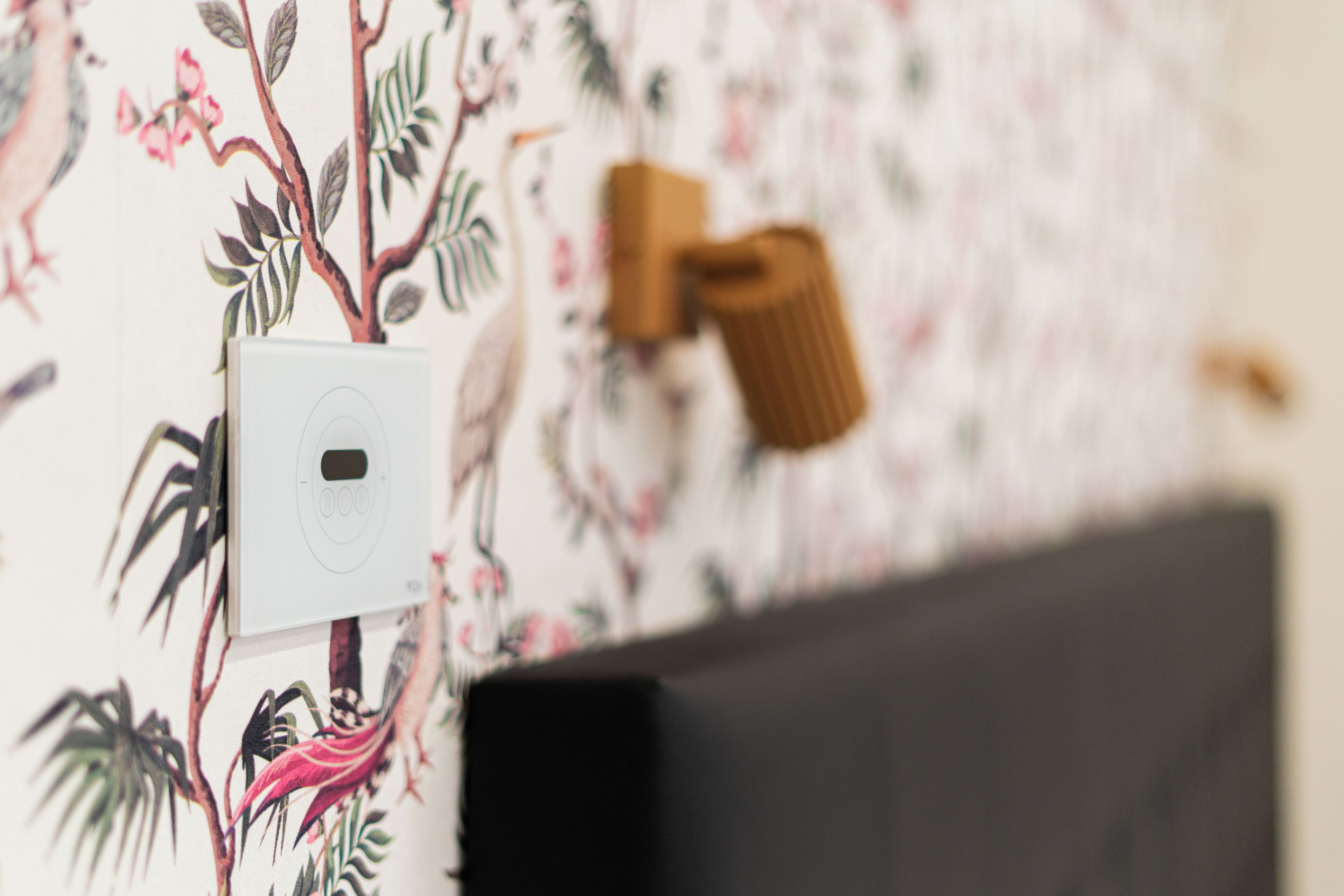 Una pared con un estampado floral, un cuadro eléctrico blanco y un objeto marrón.