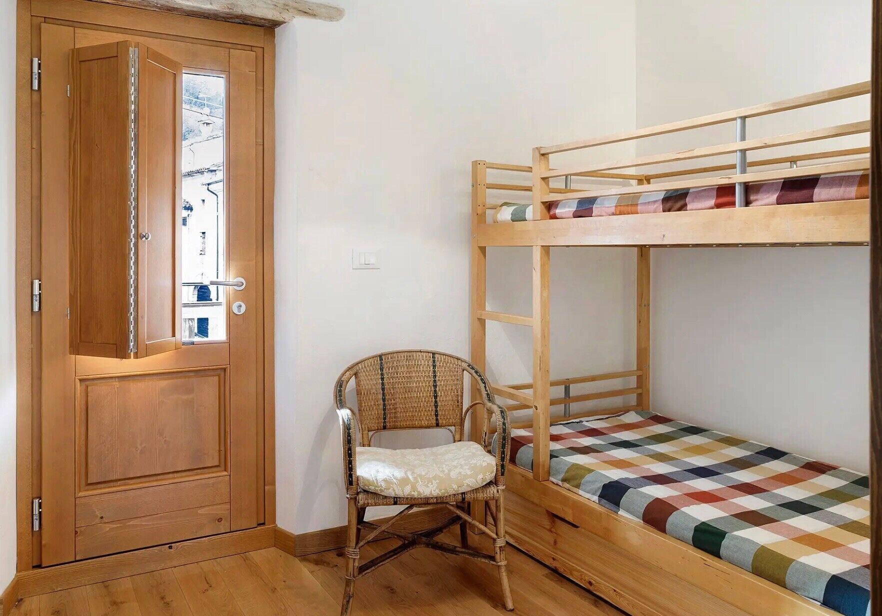 A bunk bed with a checkered bedspread, a wicker chair, and a wooden door with a view of the outside.